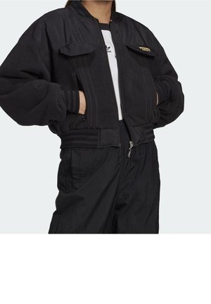 Persoon draagt een zwart bomberjack met rits en klepzakken, gecombineerd met een zwarte broek en een wit Adidas-shirt dat eronder zichtbaar is.