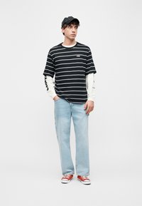 Νέος άνδρας που φοράει μαύρο καπέλο, μαύρη και λευκή ριγέ μπλούζα πάνω από μακρυμάνικο λευκό μπλουζάκι, ανοιχτόχρωμα τζιν και κόκκινα αθλητικά παπούτσια, στέκεται με τα χέρια στις τσέπες.