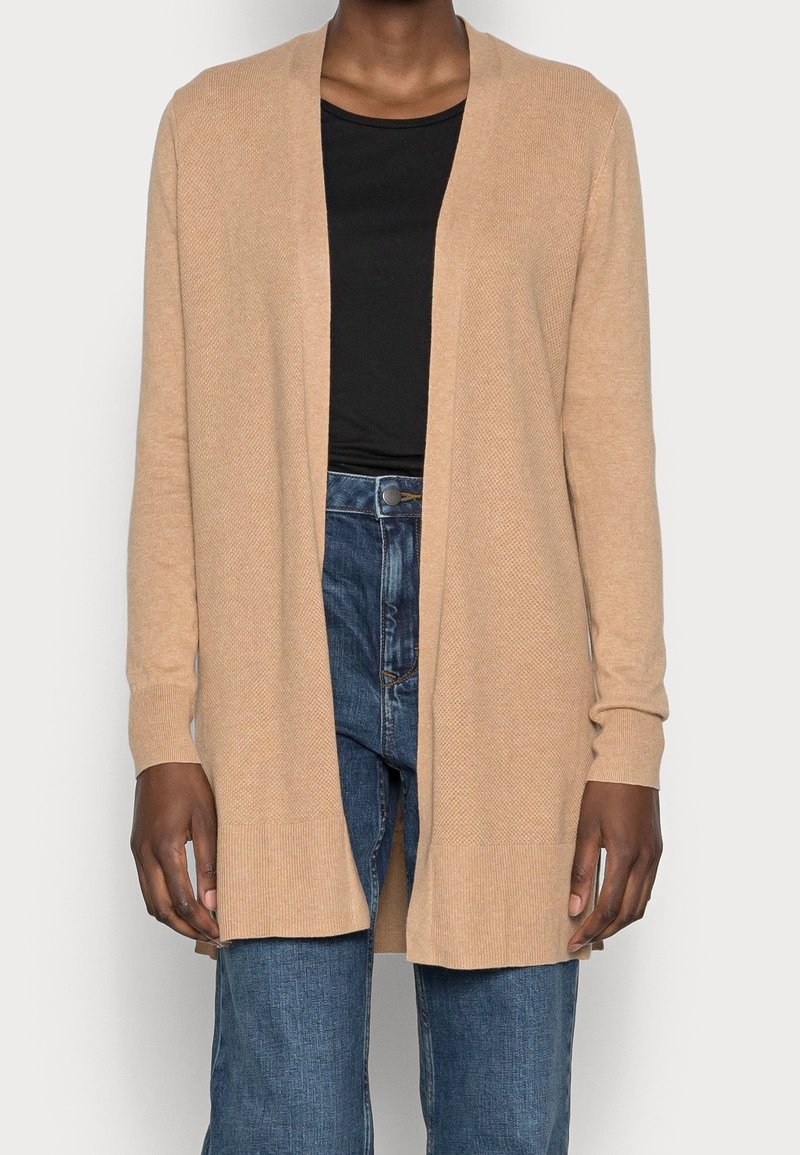 Cardigan beige en maille avec devant ouvert, manches longues et poignets côtelés. Associé à un jean bleu taille haute, affichant une coupe décontractée.