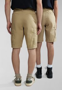 Dos personas con pantalones cortos de cargo beige y camisetas negras, vistas desde atrás, una con zapatillas deportivas y la otra con botas negras.
