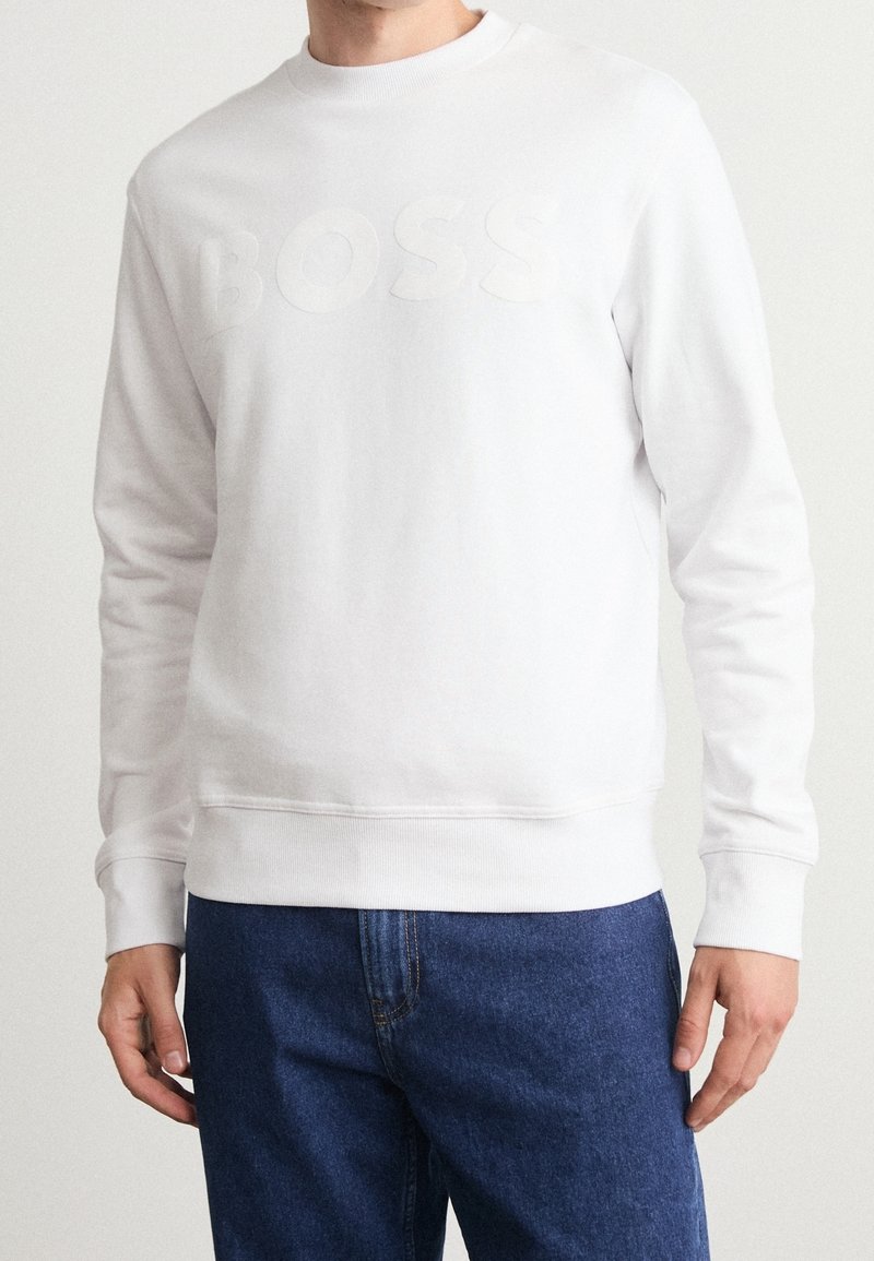 Vit crewneck-tröja med mjuk textur, med en upphöjd 'BOSS'-logotyp över bröstet. Tillsammans med blå denimjeans.