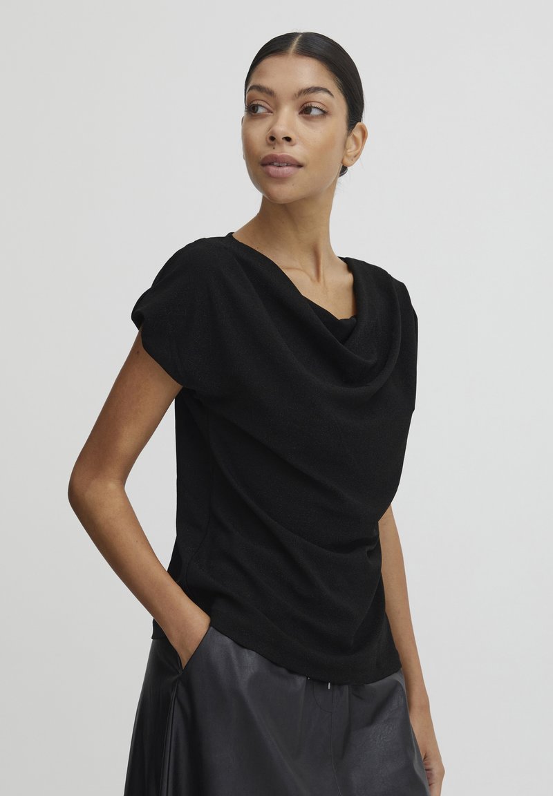 Top nero con scollo drappeggiato e maniche corte, realizzato in un tessuto testurizzato; indossato con una gonna in pelle.