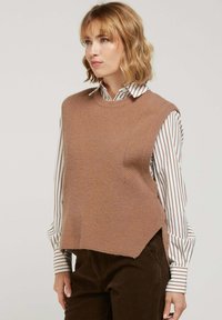 Pull en tricot sans manches couleur brun clair avec une surface texturée, doté d'un col rond et de fentes latérales, superposé sur une chemise rayée.