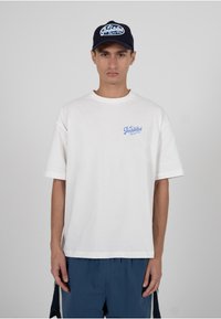 Valge puuvillane lühikeste varrukatega T-särk, millel on rinnal sinine kiri; kantud koos laigulise sinise pesapalliämbriga, millel on sarnane kiri.