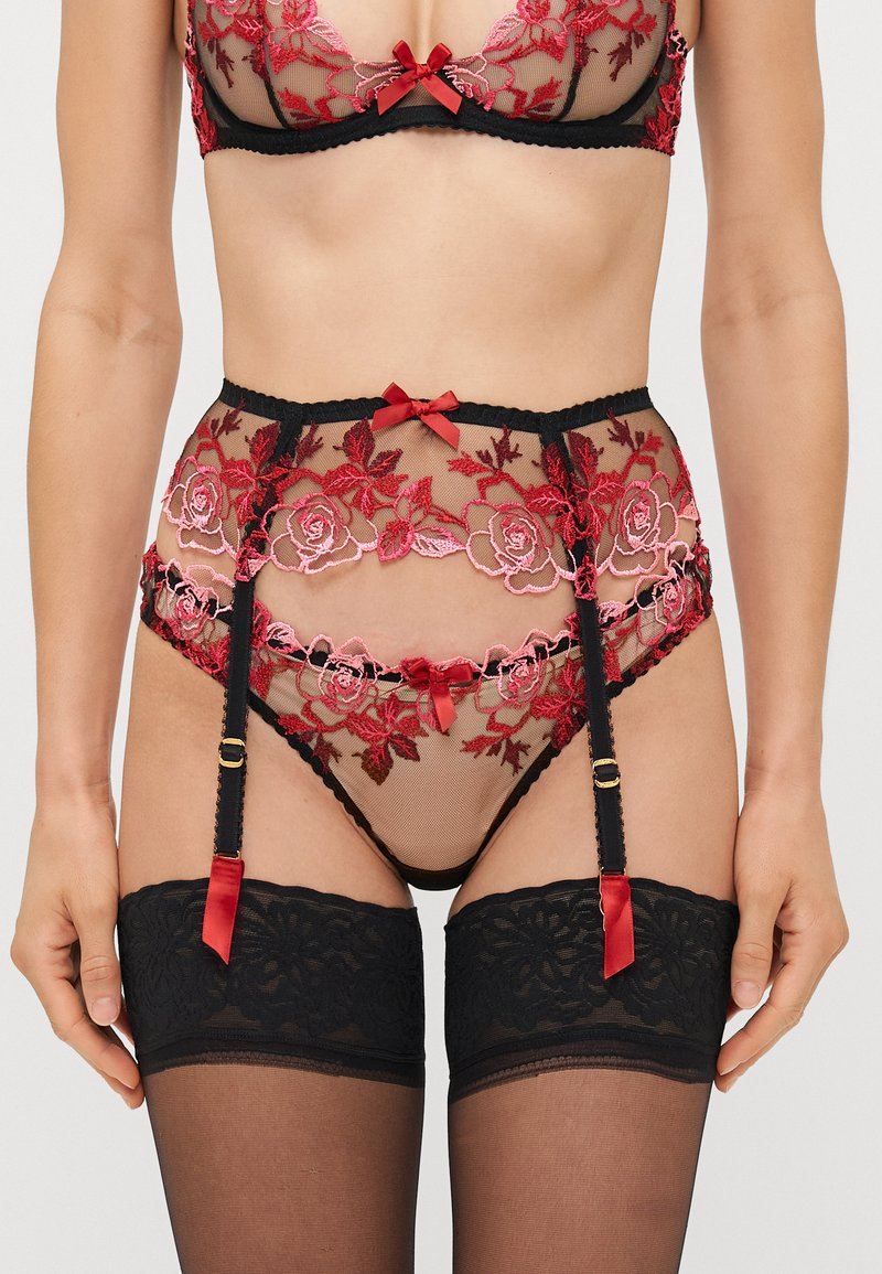Ensemble de lingerie en dentelle noire comprenant un mesh transparent avec des broderies rouges. Comprend une ceinture porte-jarretelles avec des nœuds et des bas collants assortis.