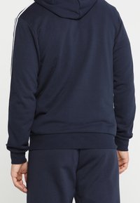 Osoba oblečená v tmavě modré mikině s kapucí a odpovídajících teplácích, stojící zády ke kameře, ruce volně podél těla.