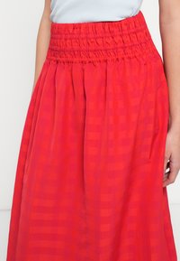 Czerwona, wzorzysta długa spódnica maxi z marszczoną gumą w pasie, wykonana z lekkiego materiału. Charakteryzuje się zwiewnym krojem i teksturą z delikatnym wzorem w kratkę.