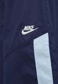 Námornícky vetrovka s svetlomodrým bočným pruhom a bielym logom Nike. Vyrobená z mäkkého, ľahkého materiálu s hladkým povrchom.