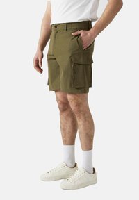 Olijfgroene cargoshorts met een elastische tailleband, zijzakken en duurzaam materiaal. Draagt met witte sneakers en korte sokken.