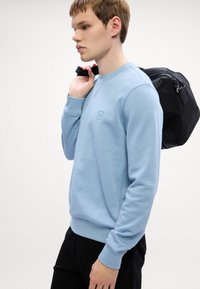 Φωτεινό μπλε φούτερ με στρογγυλή λαιμόκοψη, ριγέ μανσέτες και μικρή ετικέτα λογότυπου, φορεμένο από άνδρα μοντέλο, συνδυασμένο με μια μαύρη τσάντα.