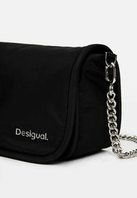 Svart nylonaxelväska med vikbar klaff, silverkedjestropp och präglad "Desigual."-logotyp på framsidan. Slät textur och minimalistisk design.