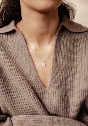 Woman wearing a silver necklace with a teardrop-shaped pendant over a textured brown knit sweater with a V-neck collar.