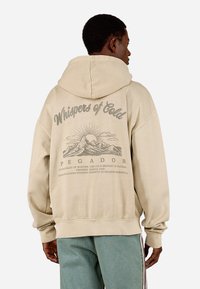 Sweatshirt oversized bege com capuz ajustável. Apresenta uma impressão gráfica de montanhas e sol nas costas. Punhos e bainha com ribana.