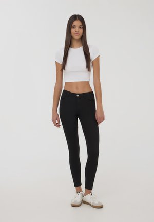 Leggings neri a vestibilità slim, con tasche frontali e chiusura con bottone anteriore, abbinati a una t-shirt corta bianca e scarpe da ginnastica bianche.