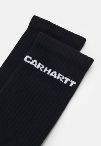 Μαύρες ριμπ κάλτσες με λευκό λογότυπο "CARHARTT" υφαντό κοντά στην κορυφή. Διαθέτουν μαλακή υφή και παραδοσιακό σχεδιασμό σε μήκος κώνου.
