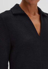 Schwarzes Strickkleid mit tiefem V-Ausschnitt und Kragen. Glatte Textur mit figurnahem Schnitt, der den Ausschnitt und den Brustbereich betont.