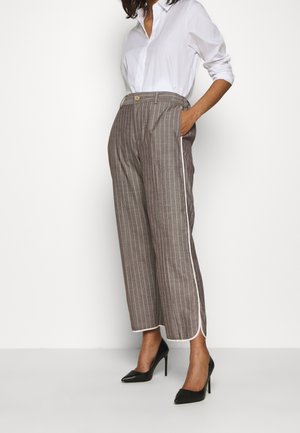 Femme portant une chemise blanche boutonnée, un pantalon large à rayures marron avec une bordure blanche sur le côté, et des chaussures à talons noirs, debout avec une main dans la poche.