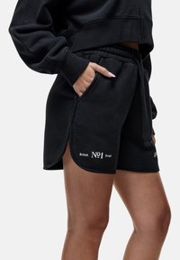 Pantaloni corti neri in cotone con spacco laterale, elastico in vita e tasche. Presentano la scritta "British No1 Design" in bianco sulla gamba sinistra.