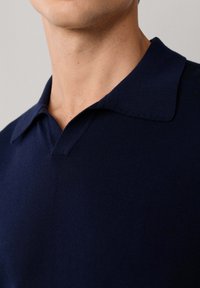 Primer plano de una persona que lleva una camisa tipo polo de punto azul marino con un cuello suave y una abertura en forma de V sutil.
