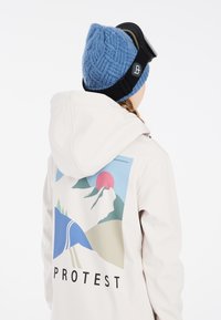 Crème-kleurige jas met een kleurrijke bergafbeelding op de achterkant, gecombineerd met een blauwe gebreide beanie en zwarte skibril.