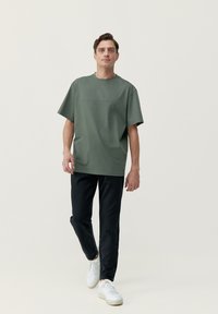 Olivovo zelené bavlnené tričko s voľným strihom, vybavené krátkymi rukávmi a jednoduchou potlačou textu na hrudi. V kombinácii s čiernymi užšími nohavicami a bielymi teniskami.