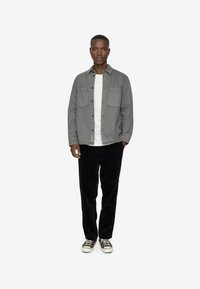 Camicia grigia a maniche lunghe con due tasche sul petto sopra una maglietta bianca, abbinata a pantaloni in velluto a coste neri e sneaker marroni. Design semplice, senza motivi.