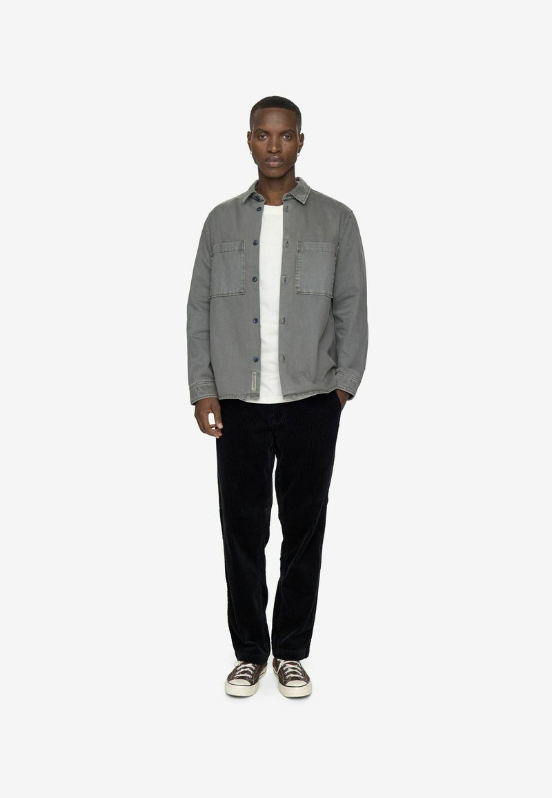 Camicia grigia a maniche lunghe con due tasche sul petto sopra una maglietta bianca, abbinata a pantaloni in velluto a coste neri e sneaker marroni. Design semplice, senza motivi.