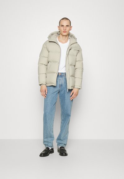 Lys grå pufferjakke med hætte, lynlås og syede sektioner. Båret over en hvid t-shirt og blå jeans, parret med sorte sko.