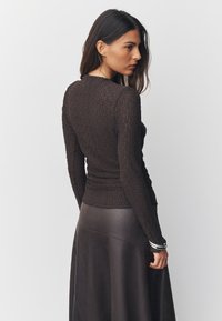 Haut marron texturé à manches longues avec un design froncé, associé à une jupe en cuir foncé et un bracelet argenté. Fond uni.