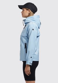 Veste imperméable bleu clair avec capuche, fermetures éclair et poignets ajustables. Présente un design ample et des poches à l'avant. Accents noirs sur les fermetures éclair.