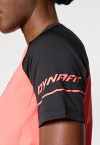 Camisa atlética de manga curta com parte superior preta e corpo em coral, com riscas vermelhas contrastantes e logótipo "DYNAFIT" na manga.