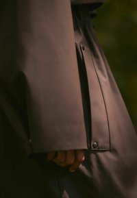 Giacca impermeabile marrone con una texture liscia, polsini aderenti e dettaglio con bottoni a pressione sulla manica, che stringe parzialmente una mano.