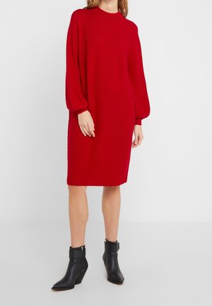Femme portant une robe rouge mi-longue avec de longues manches bouffantes et des bottines noires à talons carrés, debout devant un fond blanc.