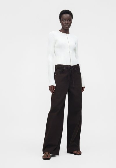 Γυναίκα που φοράει λευκό ριμπ μπλουζάκι με φερμουάρ, φαρδιά μαύρη παντελόνα με κορδόνι στη μέση και μαύρα σαγιονάρες, στέκεται σε λευκό φόντο.