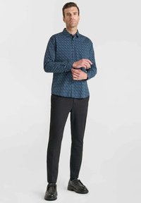 Man staat in een blauw patroon langemouwen shirt, zwarte slim-fit broek en zwarte nette schoenen voor een eenvoudige witte achtergrond.