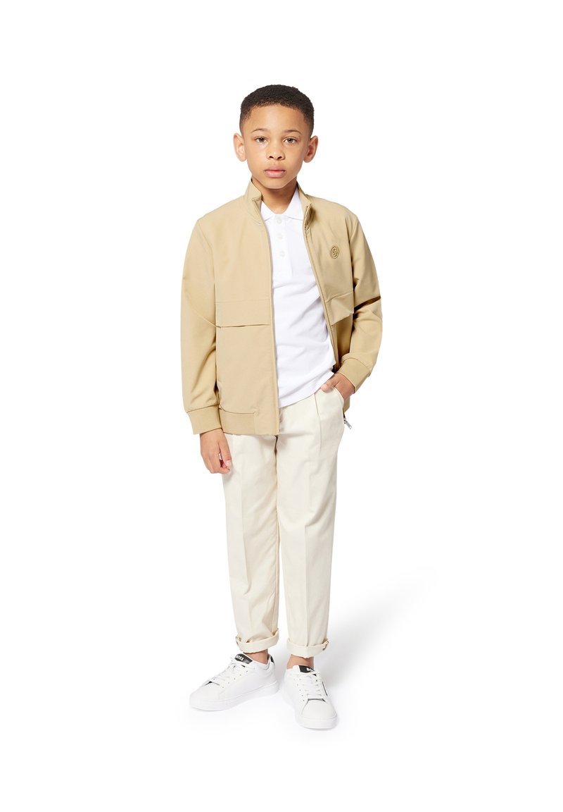Veste beige à col montant, polo blanc, pantalon crème et baskets blanches. Texture lisse, design décontracté et manches de pantalon retroussées.