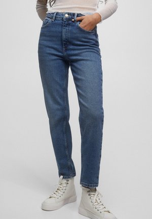 Frau trägt hoch taillierte blaue Jeans und weiße High-Top-Sneaker, mit einer Hand in der Tasche vor einfarbigem Hintergrund.