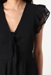 Black dress with a deep V-neck, tie detail at the front, and sheer ruffle sleeves. The fabric appears smooth with a flowing silhouette.