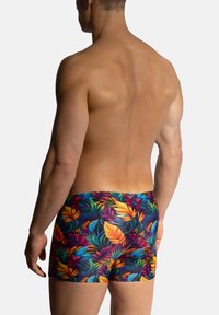 Simshorts i ett livfullt tropiskt lövmönster med flerfärgade blad på en mörk bakgrund; elastisk tyg, figurnära design.
