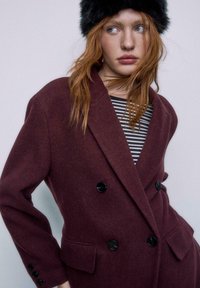 Bordeaux dobbeltknappet frakke med store sorte knapper, sidelommer og struktureret finish, parret med en sort pelshat og stribet top.