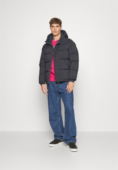 Black puffer jacket with hood, featuring quilted design, worn over a bright pink sweatshirt and blue jeans, paired with black sneakers.