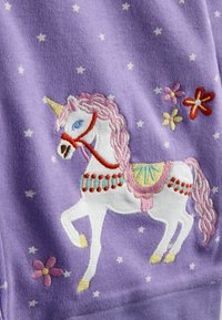 Balts vienradzis ar rozā krēpēm, dzeltenu ragu un dekoratīvu seglu, kas izšūts uz purpura auduma ar mazām baltām zvaigznītēm un krāsainiem ziediem.