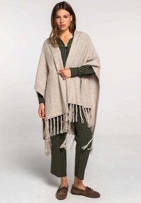 Poncho de punto beige con flecos, combinado con una camisa a cuadros en verde oscuro y negro y pantalones a juego. Zapatos mocasines marrones.