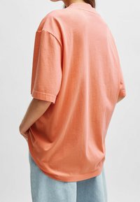 Camiseta de corte holgado y oversized en algodón color coral. Manga corta, cuello redondo, textura suave y dobladillo recto. Jeans azul claro visibles en la parte inferior.