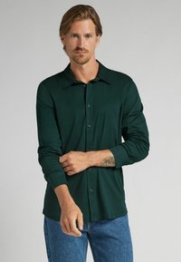 Hombre con cabello castaño claro que lleva una camisa de botones verde oscuro y jeans azules, ajustando su manga izquierda, de pie contra un fondo liso.