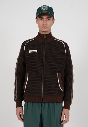 Brun strikkejakke med glidelås, teksturert mønster, kremfargede detaljer, høy krage og sidelommer. Kombinert med grønne bukser og en caps.