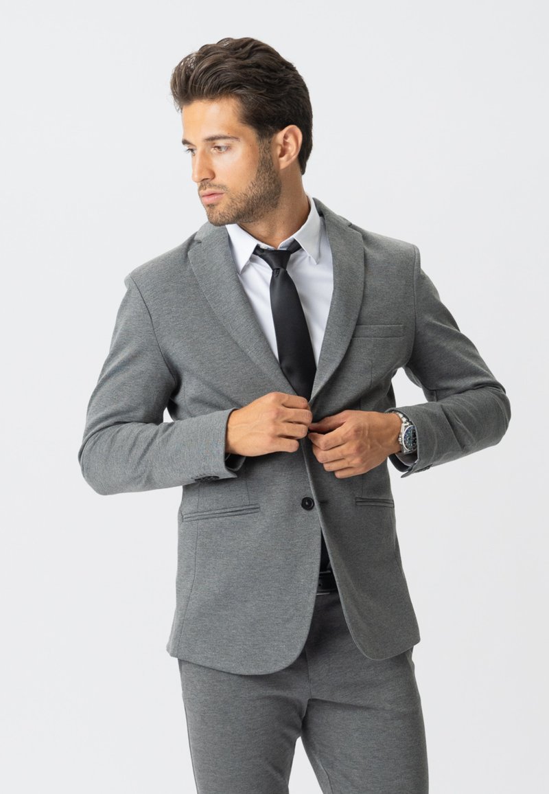 Homme boutonnant une veste de costume grise sur une chemise blanche et une cravate noire, se tenant devant un fond clair uni.