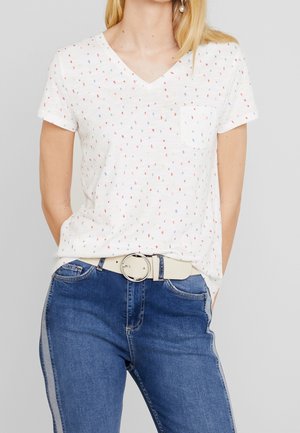 Biała koszulka z dekoltem w serek z małymi wielokolorowymi kropkami, kieszonką na piersi, zestawiona z niebieskimi dżinsami ozdobionymi jasnymi paskami po bokach oraz beżowym paskiem z okrągłą klamrą.