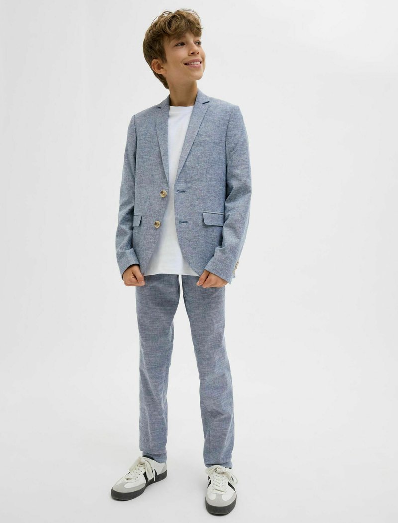 Világoskék öltöny, texturált felülettel, egysoros zakóval, két gombbal és hozzá illő nadrággal. Fehér pólóval és sportcipővel párosítva.