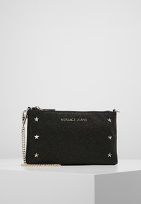 Pochette noire pailletée avec des accents d'étoiles argentées, dotée d'une fermeture éclair et d'une bandoulière chaîne amovible. Nom de la marque "VERSACE JEANS" visible.